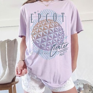 May include: A light purple t-shirt featuring a graphic of the EPCOT Center sphere with the text "EPCOT Center 1982" in a retro design. The shirt has short sleeves and a relaxed fit.