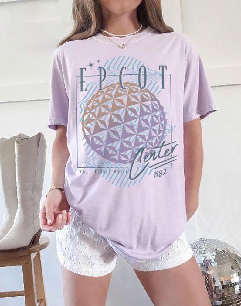 May include: A light purple t-shirt featuring a graphic of the EPCOT Center sphere with the text "EPCOT Center 1982" in a retro design. The shirt has short sleeves and a relaxed fit.