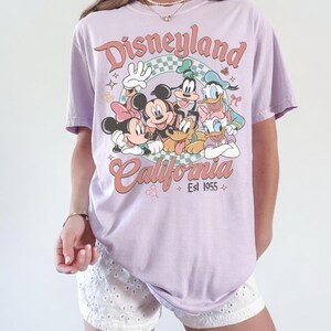 May include: A light purple t-shirt featuring a retro design with the text "Disneyland California Est 1955". The graphic includes cartoon characters Mickey Mouse, Minnie Mouse, Goofy, Donald Duck, and Pluto. The shirt has short sleeves.