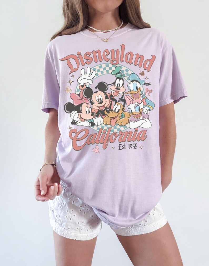 May include: A light purple t-shirt featuring a retro design with the text "Disneyland California Est 1955". The graphic includes cartoon characters Mickey Mouse, Minnie Mouse, Goofy, Donald Duck, and Pluto. The shirt has short sleeves.