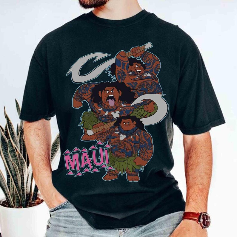 K&ouml;nnte beinhalten: Ein schwarzes Kurzarm-T-Shirt mit einem Grafikdruck der Figur Maui aus dem Film Moana in drei verschiedenen Posen. Die Figur ist mit blauen Stammes-Tattoos, einem gr&uuml;nen Bl&auml;tterrock und einem gro&szlig;en Haken dargestellt. Der Text MAUI ist in Pink gedruckt.