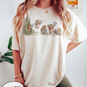 May include: Ivory-colored t-shirt featuring a whimsical illustration of Ewoks in a forest setting. The graphic depicts several Ewoks interacting with a Stormtrooper helmet and other objects. The shirt has a relaxed fit and short sleeves.