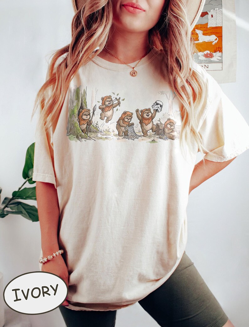 May include: Ivory-colored t-shirt featuring a whimsical illustration of Ewoks in a forest setting. The graphic depicts several Ewoks interacting with a Stormtrooper helmet and other objects. The shirt has a relaxed fit and short sleeves.