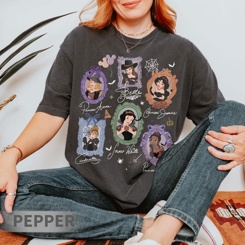 May include: Dark gray t-shirt featuring framed portraits of Disney princesses, including Aurora, Belle, Jasmine, Snow White, Cinderella, and Tiana. Each portrait is surrounded by decorative elements like bats, spiderwebs, and balloons. The princesses' names are written below their portraits.