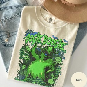 Op de afbeelding: Ivoor kleurig T-shirt met een groene afbeelding van Oogie Boogie uit The Nightmare Before Christmas. Het ontwerp bevat de tekst "Oogie Boogie" en "I am the shadow on the moon at night."