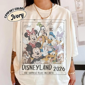 May include: Ivory-coloured t-shirt featuring a Disneyland 2026 graphic with Mickey Mouse, Minnie Mouse, Goofy, Donald Duck, Daisy Duck, Chip, and Dale. The shirt has the words "COMFORT COLORS" and "Ivory" printed on it. The text "THE HAPPIEST PLACE ON EARTH" is also printed on the shirt.