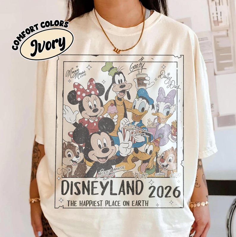 May include: Ivory-coloured t-shirt featuring a Disneyland 2026 graphic with Mickey Mouse, Minnie Mouse, Goofy, Donald Duck, Daisy Duck, Chip, and Dale. The shirt has the words "COMFORT COLORS" and "Ivory" printed on it. The text "THE HAPPIEST PLACE ON EARTH" is also printed on the shirt.