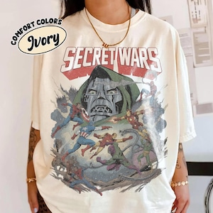 May include: An ivory t-shirt featuring a graphic print of the Marvel Secret Wars comic book cover. The design displays Doctor Doom in the background with various superheroes and villains in action, including Spider-Man, Iron Man, and Captain America.