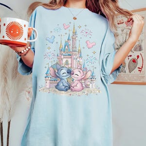May include: A light blue t-shirt featuring a graphic print of a pink and blue castle with fireworks, stars, and Mickey Mouse-shaped balloons. In the foreground, two cartoon characters, one blue and one pink, sit together in front of the castle.