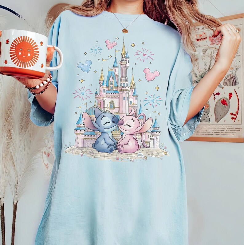 May include: A light blue t-shirt featuring a graphic print of a pink and blue castle with fireworks, stars, and Mickey Mouse-shaped balloons. In the foreground, two cartoon characters, one blue and one pink, sit together in front of the castle.