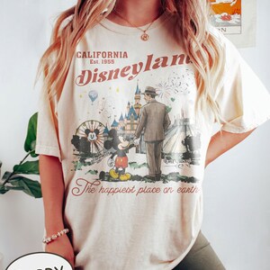 Puede incluir: Camiseta color marfil con un gr&aacute;fico vintage de Disneyland. El dise&ntilde;o incluye a Mickey Mouse, un hombre con traje, una noria y un castillo. El texto dice "California Disneyland Est. 1955" y "The happiest place on earth."