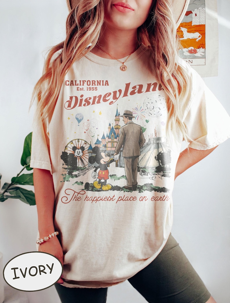 Puede incluir: Camiseta color marfil con un gr&aacute;fico vintage de Disneyland. El dise&ntilde;o incluye a Mickey Mouse, un hombre con traje, una noria y un castillo. El texto dice "California Disneyland Est. 1955" y "The happiest place on earth."