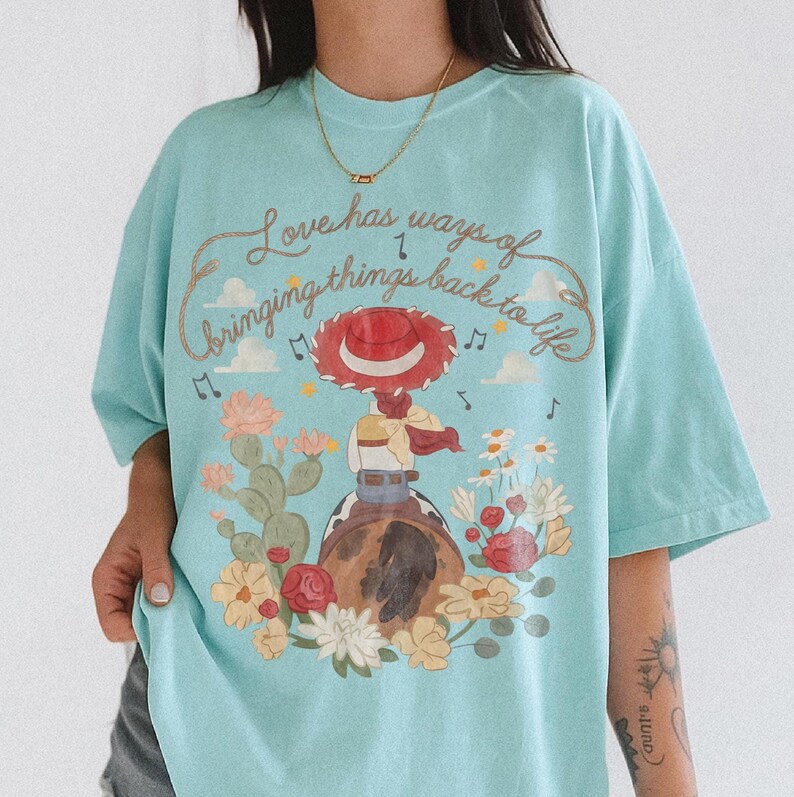 May include: A light blue t-shirt featuring a graphic of a cowgirl in a red hat riding a horse, surrounded by cacti and flowers. The text "Love has ways of bringing things back to life" is printed in a cursive font above the illustration.