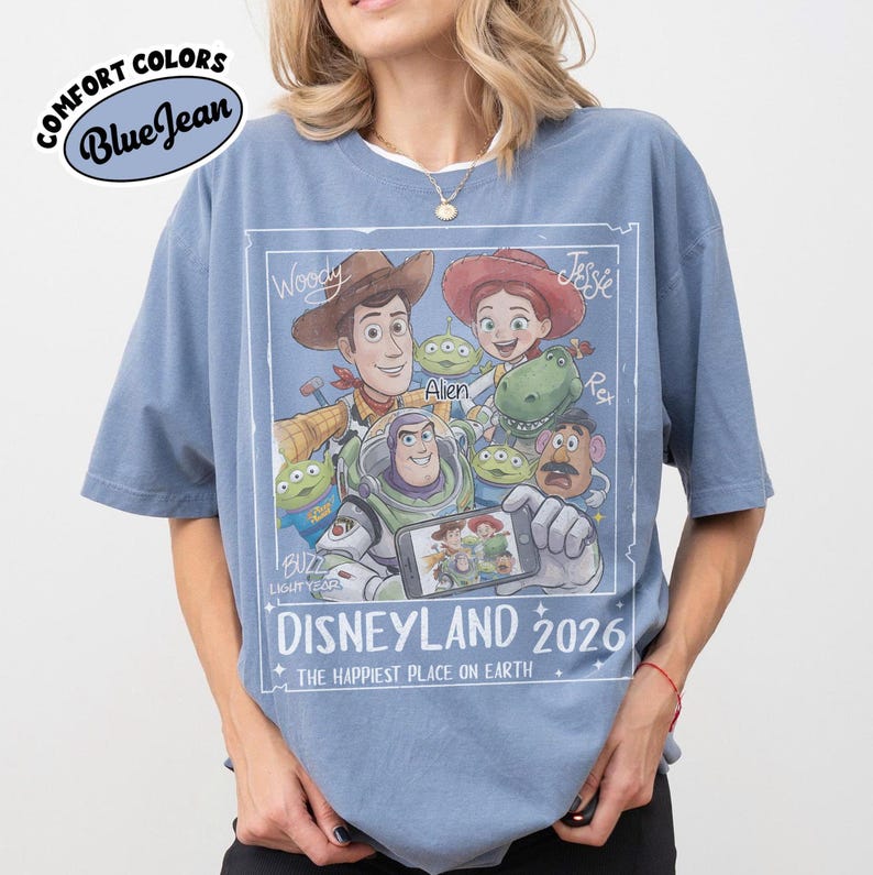 May include: Blue jean Comfort Colors t-shirt featuring a graphic of Toy Story characters, including Woody, Buzz Lightyear, and Jessie. The graphic includes the text "DISNEYLAND 2026" and "THE HAPPIEST PLACE ON EARTH".