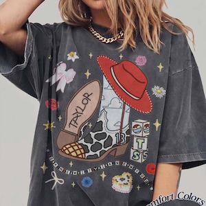 May include: A charcoal gray t-shirt featuring a graphic print of a cow-print cowboy boot, a red cowboy hat, alphabet blocks spelling TS, and the text TAYLOR and I KNEW IT I KNEW YOU. The design includes small flowers, bows, and stars.