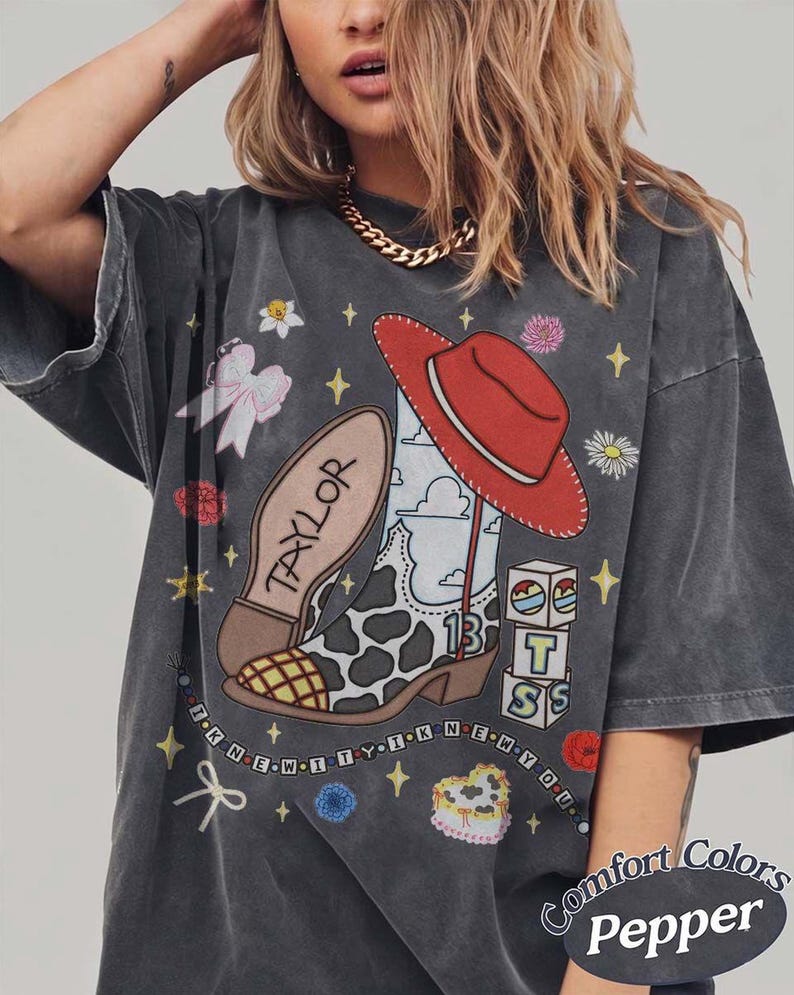 May include: A charcoal gray t-shirt featuring a graphic print of a cow-print cowboy boot, a red cowboy hat, alphabet blocks spelling TS, and the text TAYLOR and I KNEW IT I KNEW YOU. The design includes small flowers, bows, and stars.