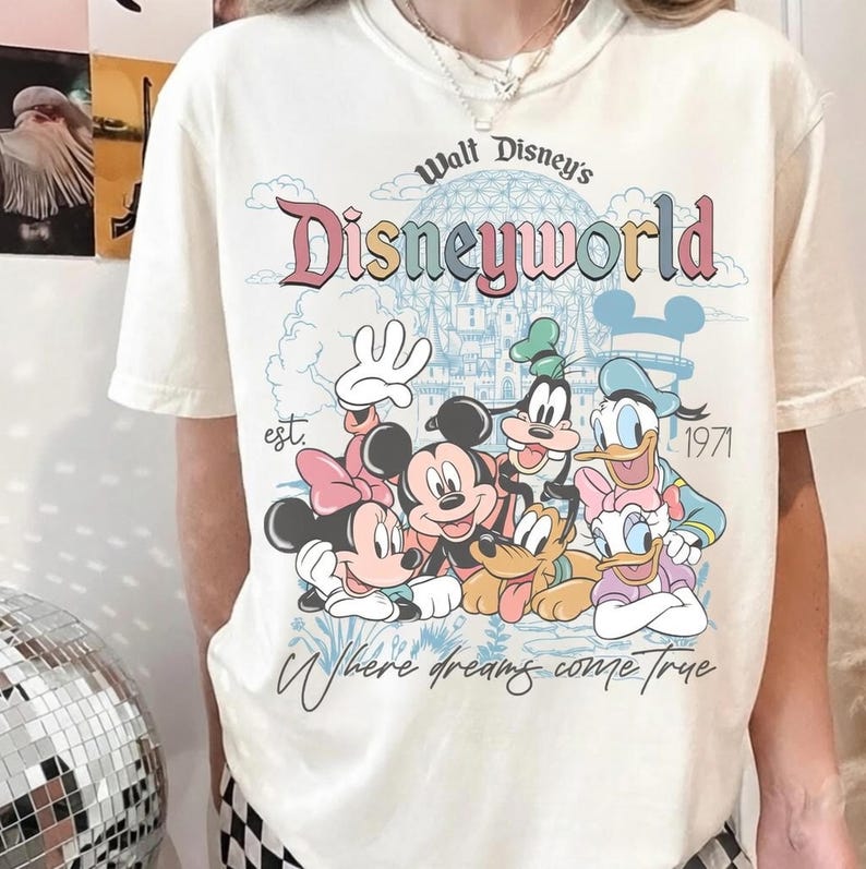 May include: Cream-colored t-shirt featuring a colorful Disney World graphic. The design includes Mickey Mouse, Minnie Mouse, Goofy, Donald Duck, and Daisy Duck. Text reads "Walt Disney's Disneyworld" and "Where dreams come true."