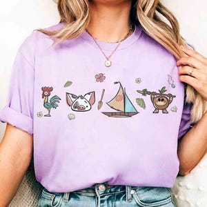Pode incluir: Uma t-shirt roxo-claro com uma estampa gr&aacute;fica de um galo, um rosto de porco, um remo de madeira, um veleiro com uma vela em espiral e um personagem de coco segurando uma lan&ccedil;a, rodeados por pequenas flores e folhas tropicais.