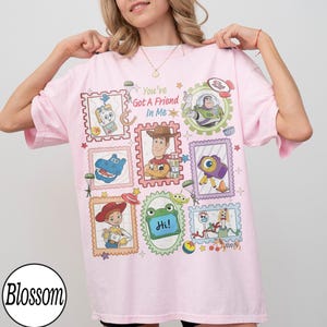 May include: A light pink t-shirt featuring a graphic print of various Toy Story characters arranged in postage stamp-style frames. The text "You've Got A Friend In Me" is printed in the centre, surrounded by illustrations of Woody, Buzz Lightyear, Jessie, Forky, and other iconic movie elements.