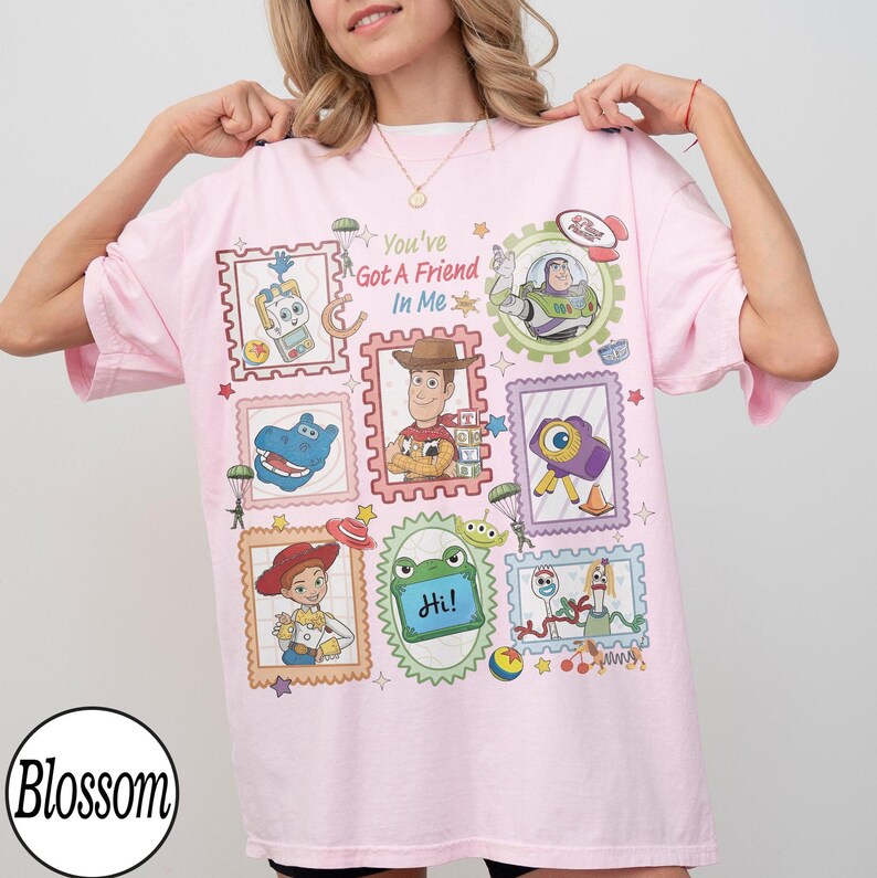 May include: A light pink t-shirt featuring a graphic print of various Toy Story characters arranged in postage stamp-style frames. The text "You've Got A Friend In Me" is printed in the centre, surrounded by illustrations of Woody, Buzz Lightyear, Jessie, Forky, and other iconic movie elements.