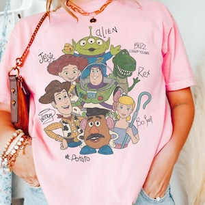May include: A light pink short-sleeve t-shirt featuring a graphic print of Toy Story characters including Woody, Buzz Lightyear, Jessie, Rex, Bo Peep, Mr. Potato Head, and an Alien, with their names written in a handwritten script style font.