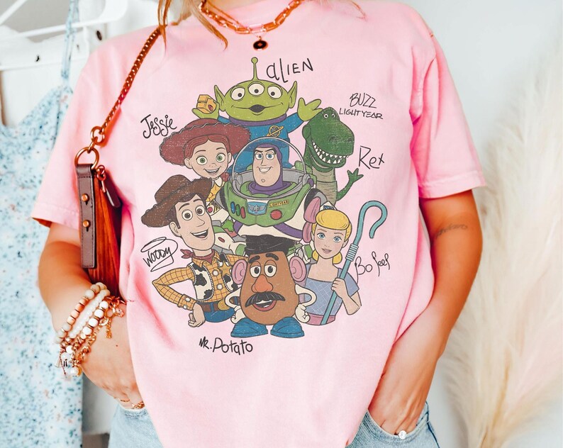 May include: A light pink short-sleeve t-shirt featuring a graphic print of Toy Story characters including Woody, Buzz Lightyear, Jessie, Rex, Bo Peep, Mr. Potato Head, and an Alien, with their names written in a handwritten script style font.