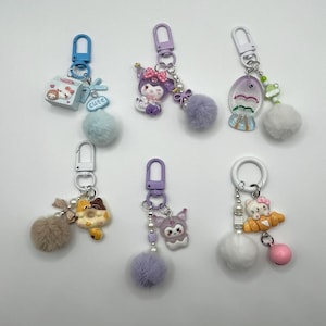 May include: Six colorful keychains with different charms, including a cartoon character, a donut, a fish, and a house. Each keychain has a fluffy pom-pom attached.