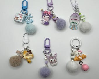 May include: Six colorful keychains with different charms, including a cartoon character, a donut, a fish, and a house. Each keychain has a fluffy pom-pom attached.
