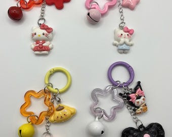 May include: Four colorful keychains with cartoon characters. Each keychain features a star-shaped charm, a bell, and a character charm. The characters include Hello Kitty, My Melody, and Pompompurin.