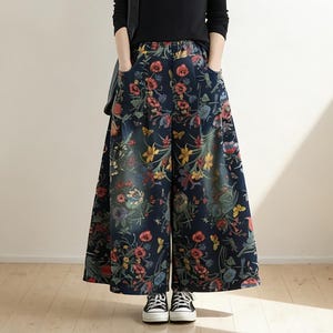 Op de afbeelding: Een persoon in een zwarte top met lange mouwen en een broek met wijde pijpen. De broek is donkerblauwe denim met een kleurrijk bloemenpatroon. De outfit wordt gecompleteerd met zwarte sneakers en een zwarte schoudertas.