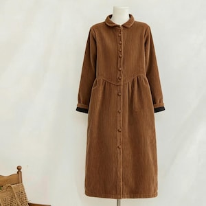 Może przedstawiać: Brązowa sztruksowa sukienka z długimi rękawami i zapięciem na guziki z przodu. Sukienka ma okrągły kołnierz i dopasowaną talię. Rękawy mają czarne mankiety. Mały drewniany fotel bujany i tkana torba są w tle.