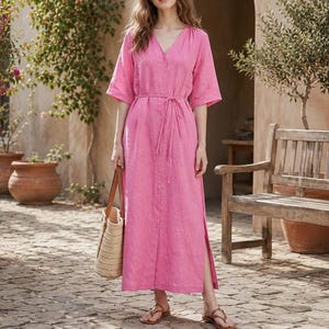 May include: A long, pink linen dress with a V-neck and short sleeves. The dress has a tie waist and side slits. A straw tote bag with a leather handle is held in the hand. The person is wearing brown sandals.