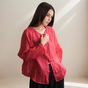 May include: A red linen blouse with long sleeves and a round neckline. The blouse has a button-down front and is paired with black pants. The fabric appears lightweight and breathable, suitable for warmer weather.