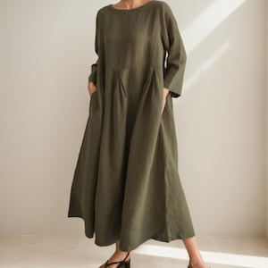 Könnte beinhalten: Olivgrünes Leinenkleid mit rundem Ausschnitt und langen Ärmeln. Das Kleid hat ein gefaltetes Oberteil und Taschen. Der Rock ist ab der Taille ausgestellt. Die Person trägt schwarze Sandalen.