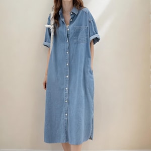 May include: A light blue denim shirt dress with short sleeves, a button-down front, and a chest pocket. The dress is knee-length and has side pockets. The model is wearing black sandals.