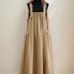 Puede incluir: Una mujer con un top negro sin mangas y un vestido con peto color beige. El vestido tiene bolsillos grandes y tirantes ajustables con hebillas metálicas. El vestido es de color liso y llega hasta los tobillos.