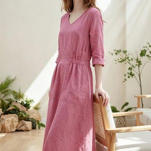 May include: A dusty rose-colored midi dress with a V-neck and three-quarter sleeves. The dress features a gathered waist, two front pockets, and a relaxed fit. The model is standing next to a wooden chair.