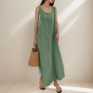 May include: A sleeveless, sage green maxi dress with a scoop neckline. The dress is made of a lightweight fabric and has a flowing silhouette. A woven straw tote bag with brown leather handles is held in the hand.