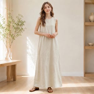 May include: A sleeveless, full-length dress in a neutral beige hue. The dress features a simple, relaxed design with a round neckline. The model is wearing brown sandals. The dress appears to be made of a lightweight fabric, suitable for warmer weather.