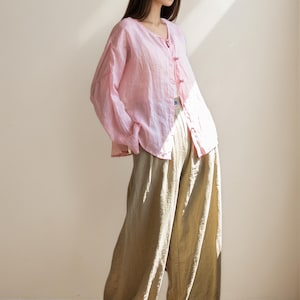 May include: A light pink linen button-down shirt with long sleeves and a V-neck, paired with wide-leg, olive-green linen pants. The outfit is loose-fitting and casual, ideal for warm weather. The shirt has small, pink buttons.