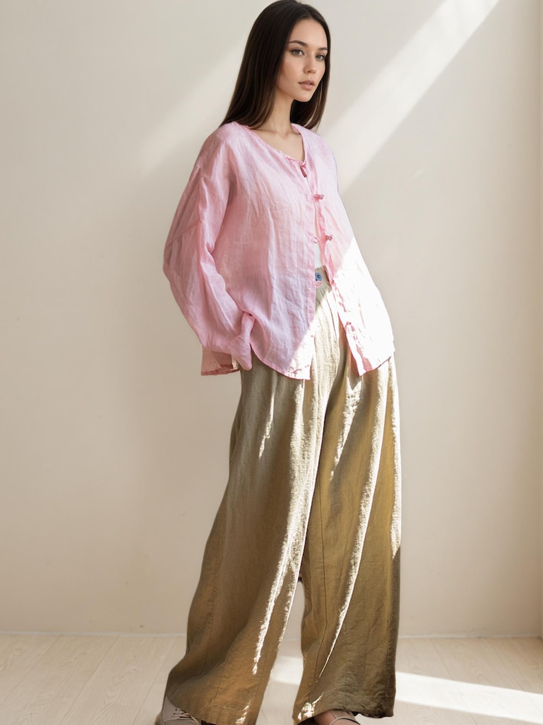 May include: A light pink linen button-down shirt with long sleeves and a V-neck, paired with wide-leg, olive-green linen pants. The outfit is loose-fitting and casual, ideal for warm weather. The shirt has small, pink buttons.