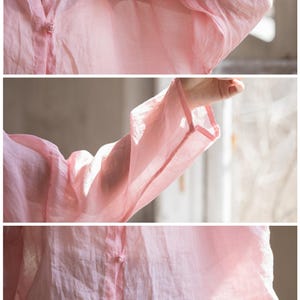 May include: A light pink, long-sleeved blouse with a V-neck and button closure. The blouse features delicate, decorative buttons and wide sleeves. The fabric appears lightweight and slightly sheer, suggesting a linen or cotton blend.