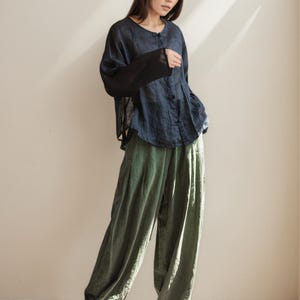 May include: A woman wearing a dark blue button-down shirt with sheer black sleeves and olive green wide-leg pants. The outfit is paired with white sneakers. The clothing appears to be made of linen or a similar lightweight fabric.