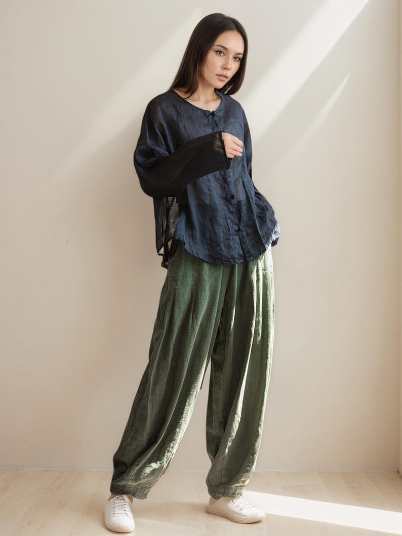 May include: A woman wearing a dark blue button-down shirt with sheer black sleeves and olive green wide-leg pants. The outfit is paired with white sneakers. The clothing appears to be made of linen or a similar lightweight fabric.