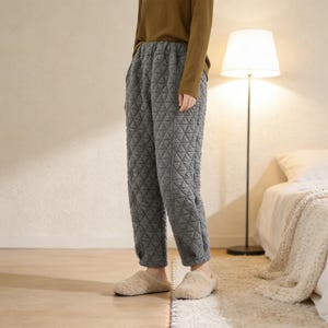 Könnte beinhalten: Graue, gesteppte Schlafanzughose mit geometrischem Muster. Die Hose hat einen elastischen Bund und gerippte Knöchel. Die Person trägt ein langärmliges braunes Oberteil und beige Hausschuhe. Die Hose scheint aus einem weichen, warmen Material zu sein.