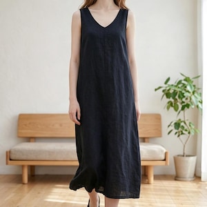 May include: A sleeveless, black V-neck dress. The dress is knee-length and made of a flowing fabric. The person is wearing black sandals. The background includes a wooden bench and a potted plant.
