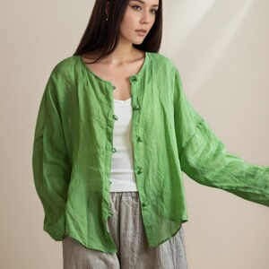 May include: A woman wearing a lime green linen button-up shirt over a white tank top, paired with gray linen pants. The shirt has long sleeves and a round neckline. The outfit is casual and comfortable.