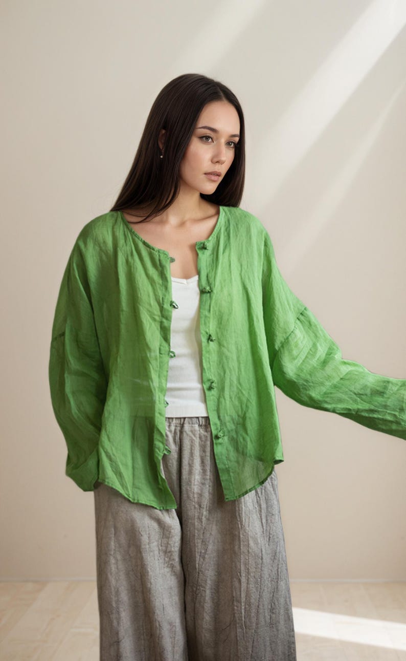 May include: A woman wearing a lime green linen button-up shirt over a white tank top, paired with gray linen pants. The shirt has long sleeves and a round neckline. The outfit is casual and comfortable.