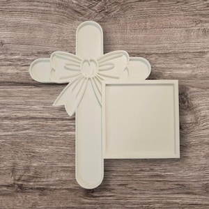 May include: A cream-colored cross-shaped photo frame with a bow detail. The frame has a square opening for a photo and is made of a light-colored material. The cross has rounded edges and is set against a wood-grain background.