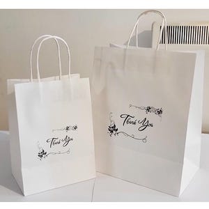 May include: Two white paper gift bags with black handles and floral designs. The larger bag has the text "Thank You" printed on it.